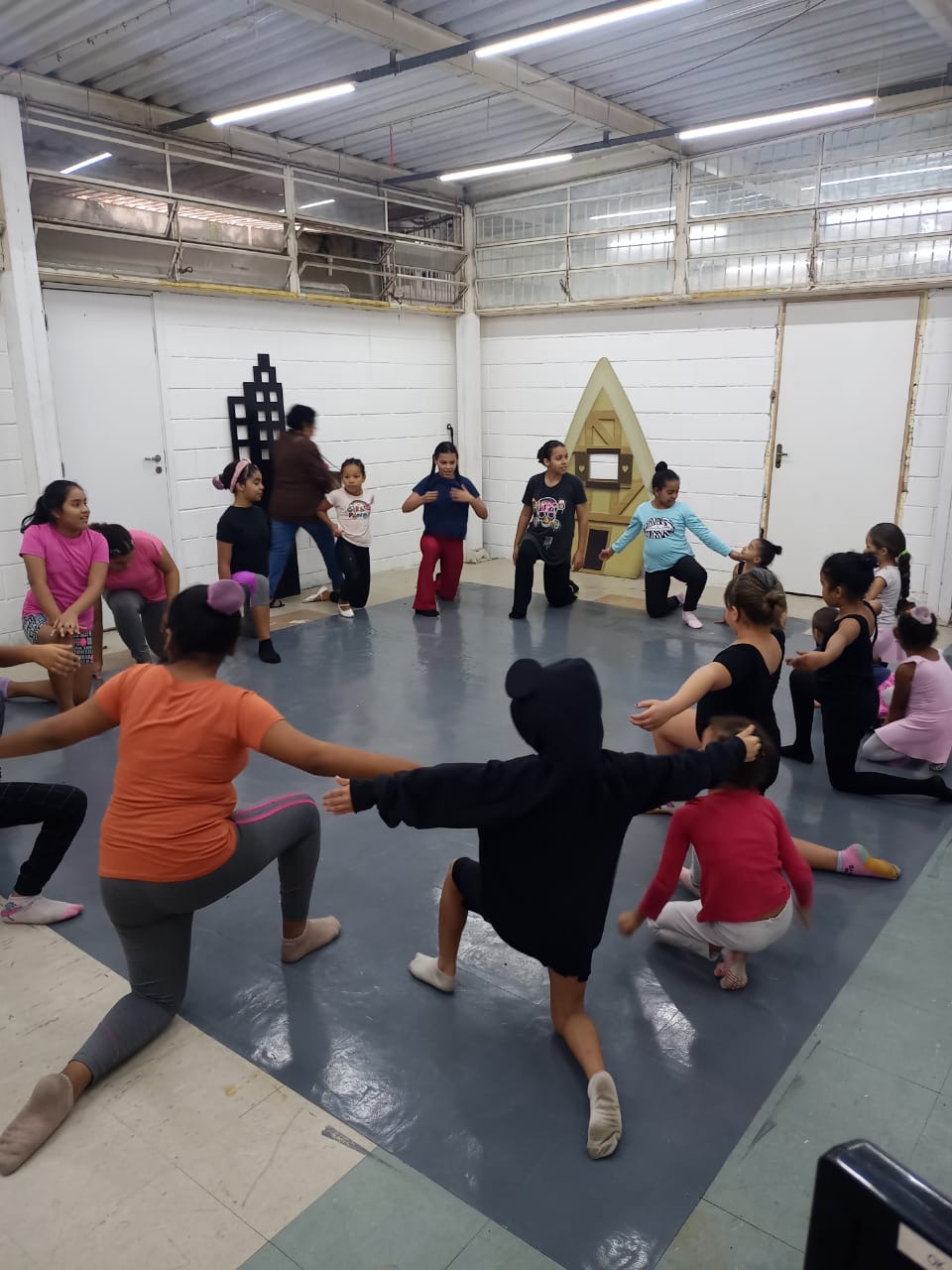 Crianças em roda durante aula de balé, acompanhadas por uma adulta ao fundo da sala