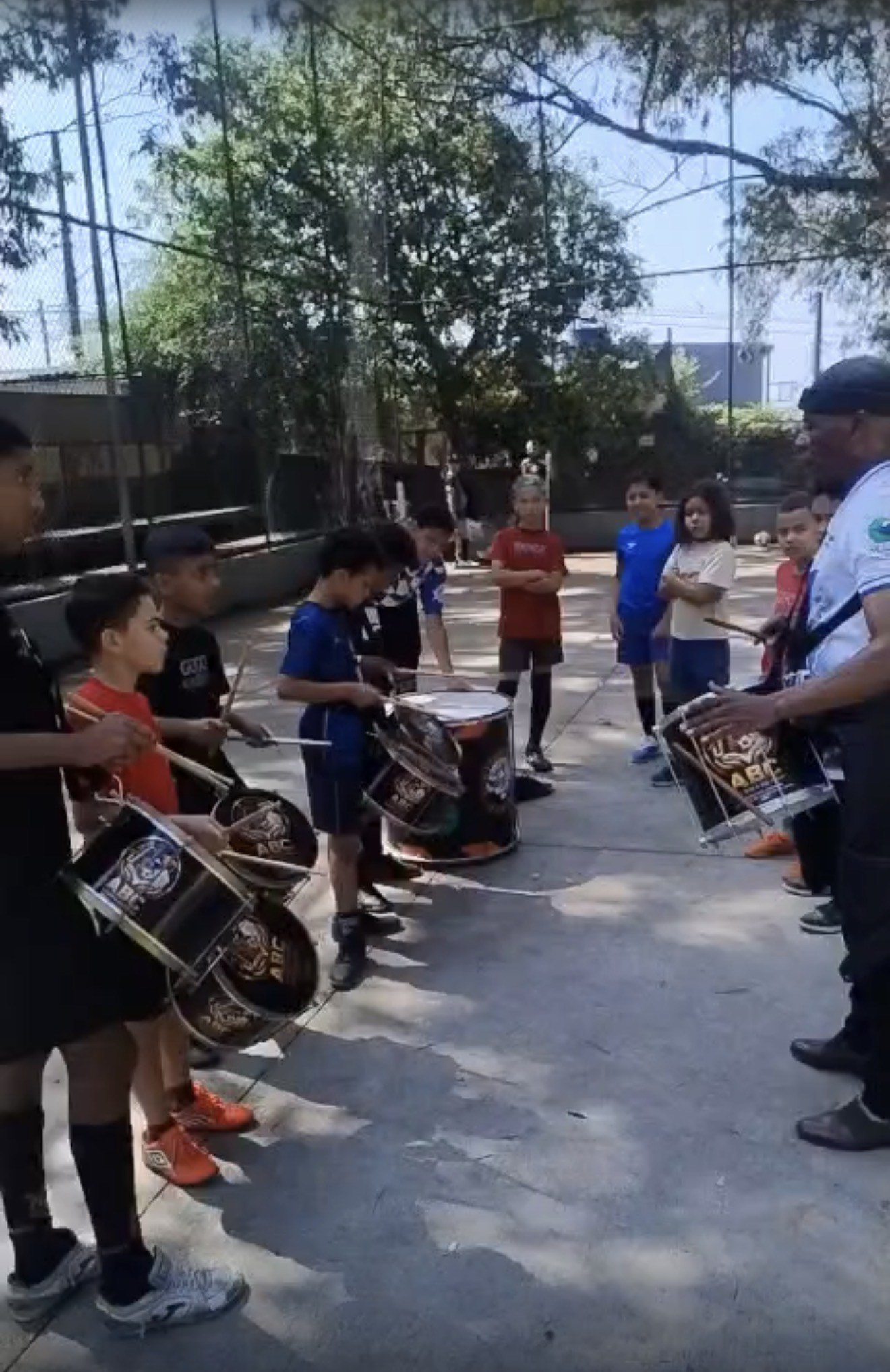 Educador orientando um grupo de crianças com caixas e surdo durante oficina de percussão em uma quadra