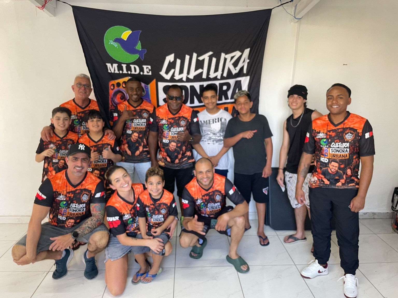 Equipe e participantes com camisetas do Cultura Sonora Urbana posando diante de um banner do MIDE e do projeto