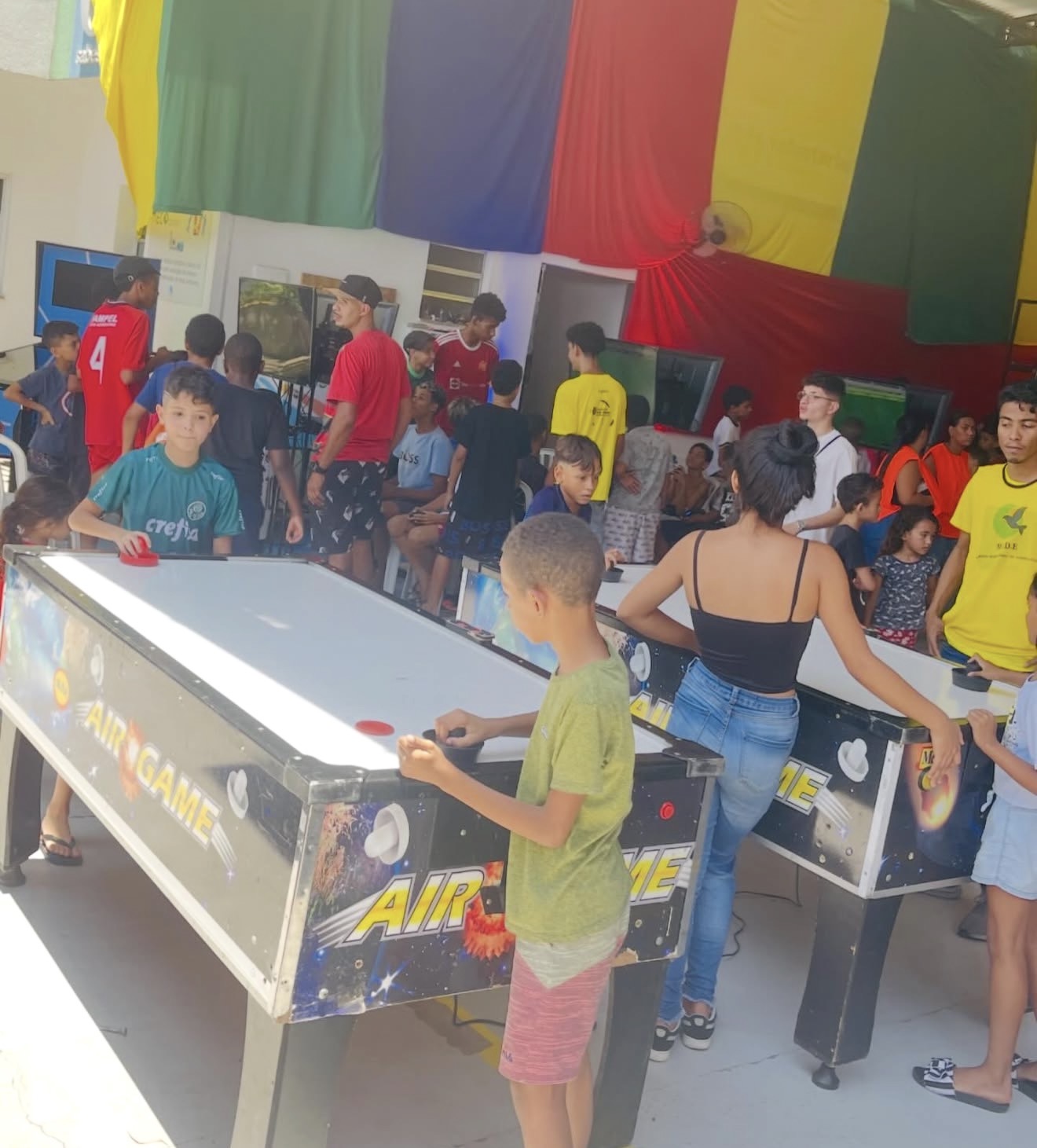 Crianças e jovens jogando em mesas de aero-hóquei em espaço coberto com tecidos coloridos