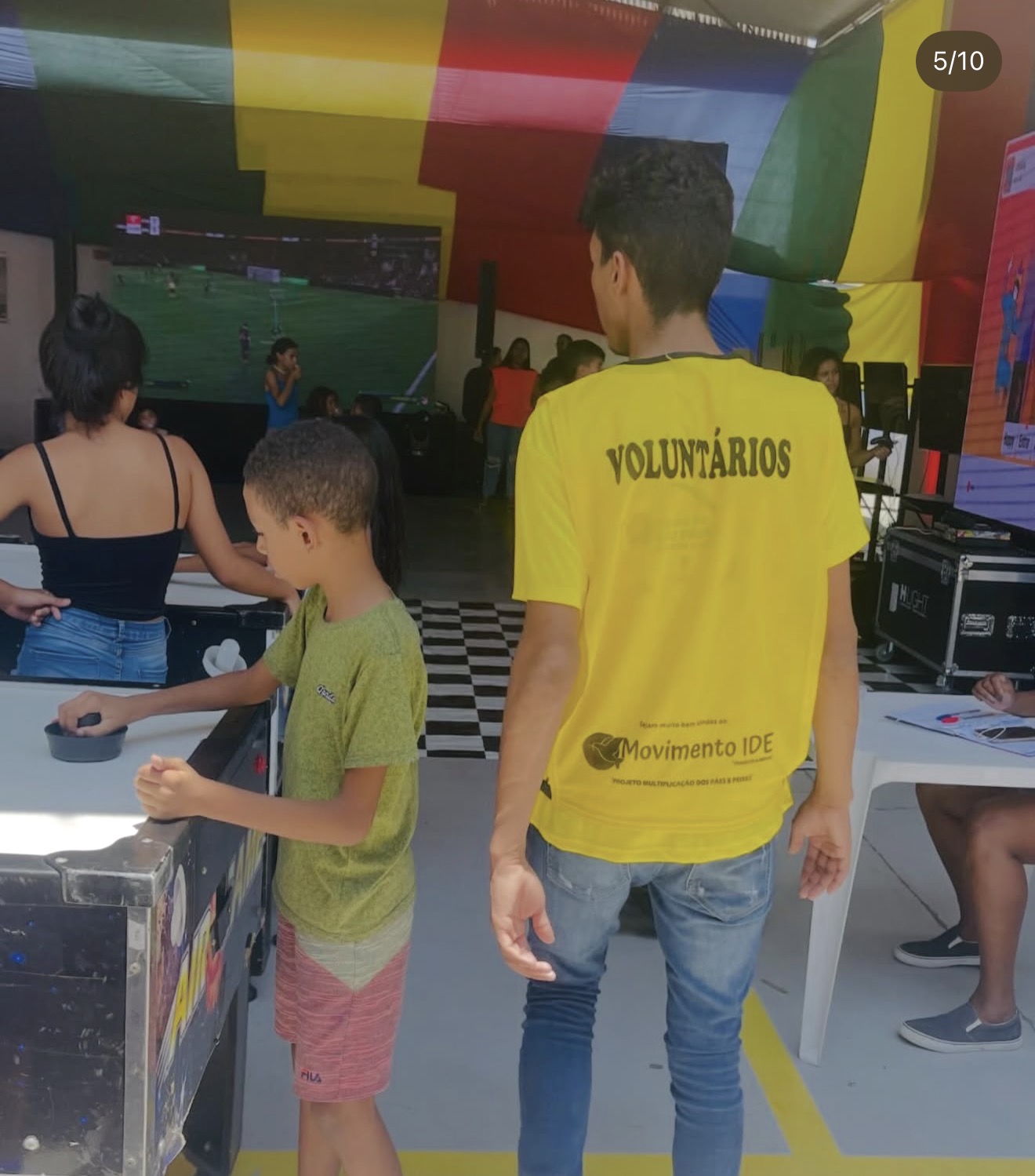 Voluntário de costas com camiseta amarela do Movimento IDE, ao lado de criança jogando aero-hóquei