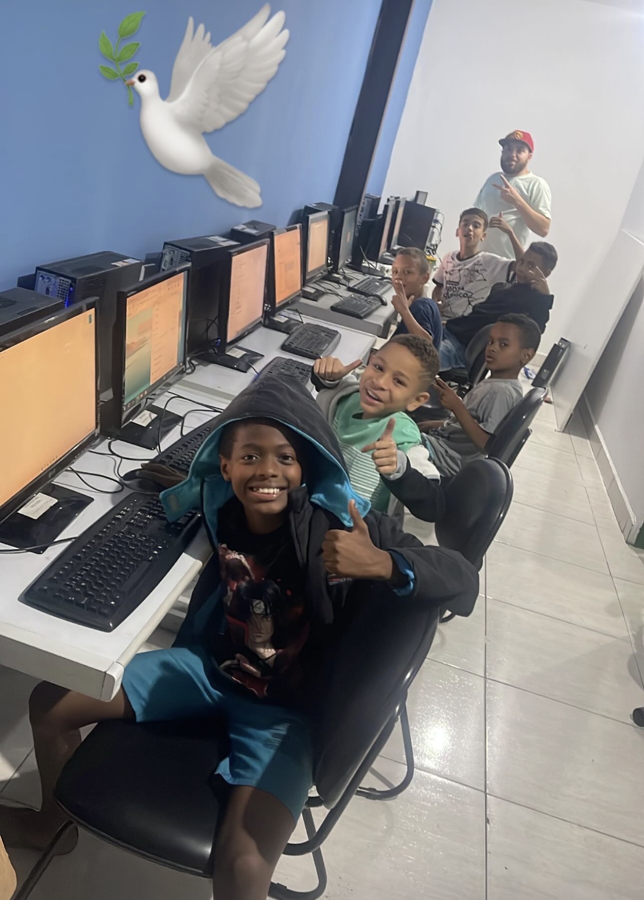 Crianças sentadas diante de computadores sorrindo para a foto, com um adulto ao fundo