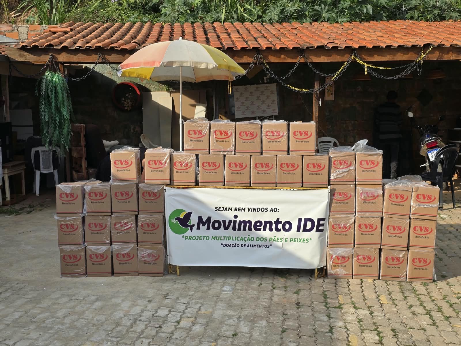 Caixas de alimentos empilhadas ao redor de uma mesa com a faixa "Sejam bem-vindos ao Movimento IDE — Projeto Multiplicação dos Pães e Peixes — Doação de alimentos"