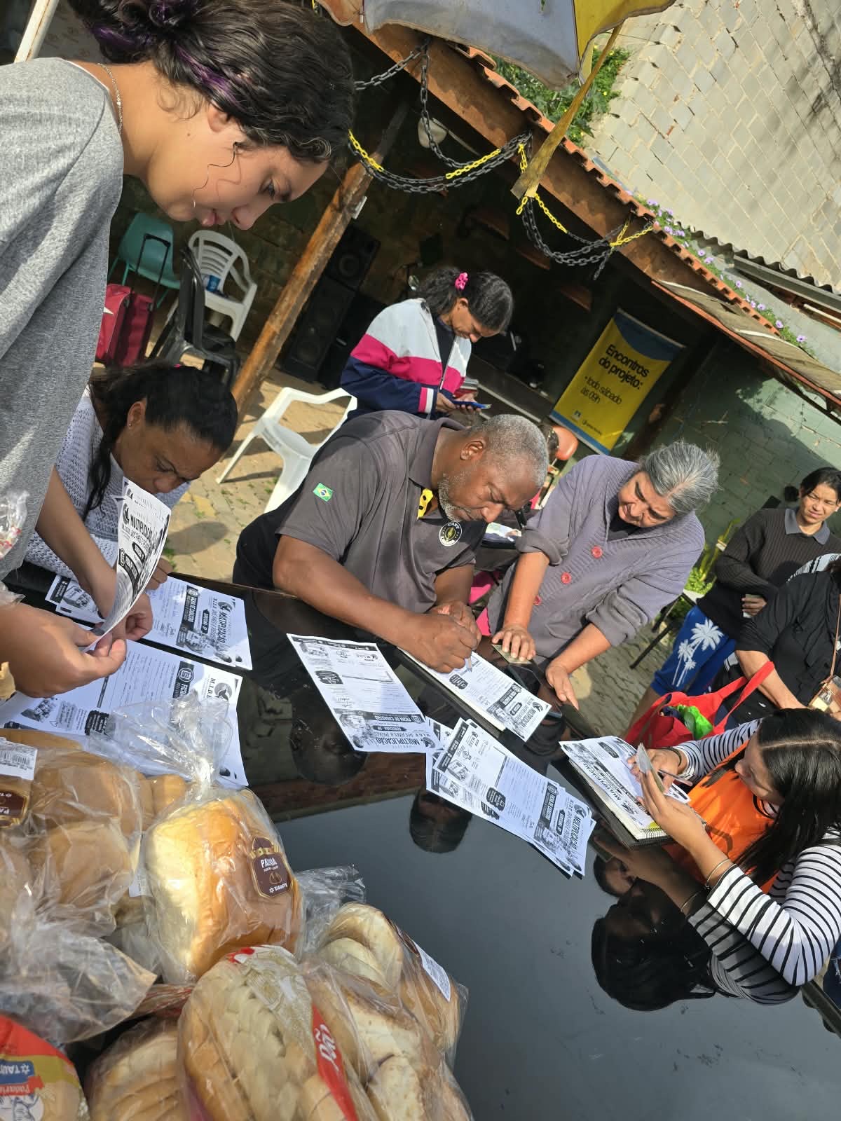 Pessoas preenchendo fichas sobre uma mesa durante o atendimento, com pães embalados em primeiro plano
