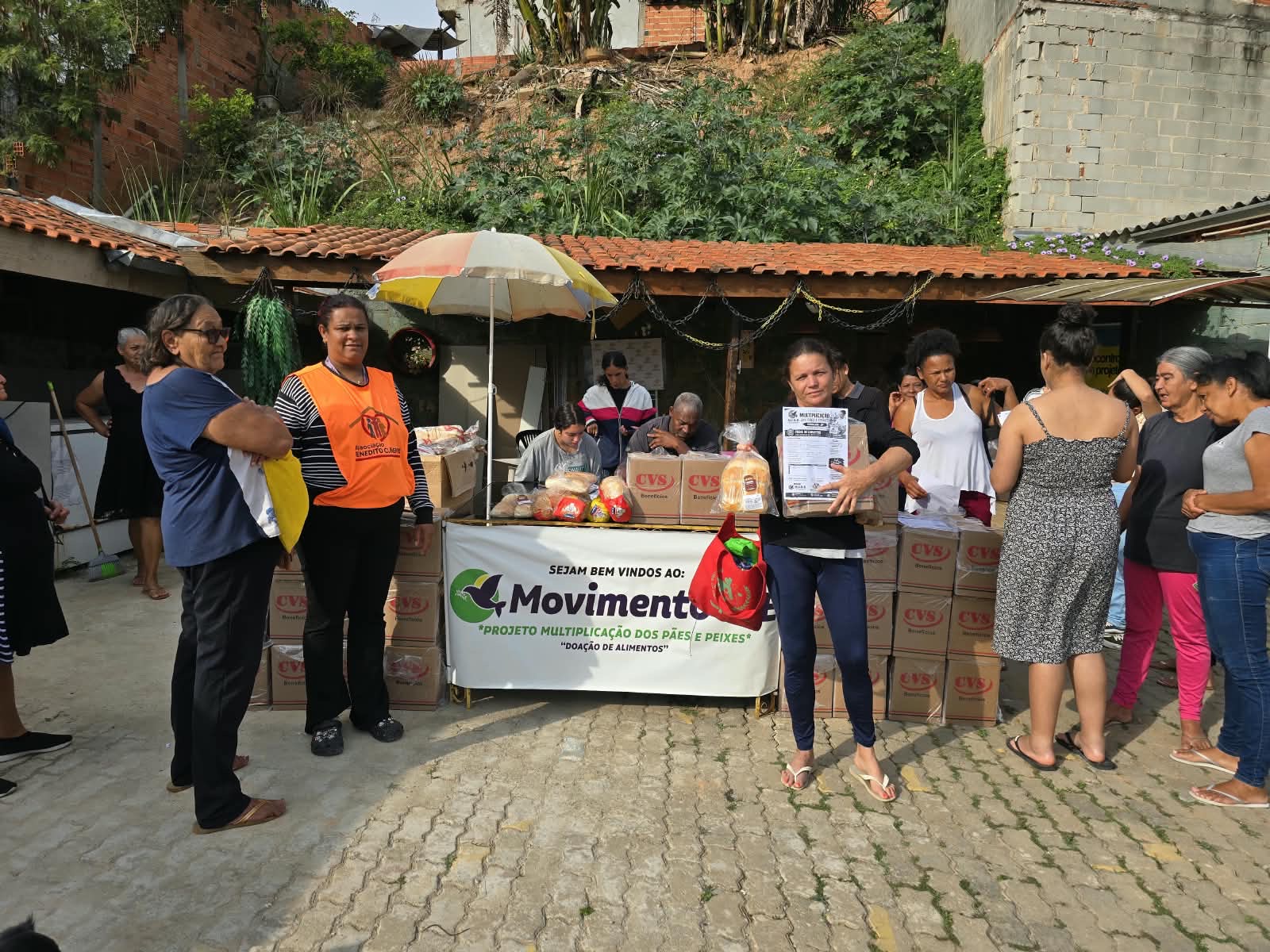 Participante segurando uma caixa de alimentos e pão ao lado da mesa do projeto, com voluntária de colete laranja e outras pessoas ao redor