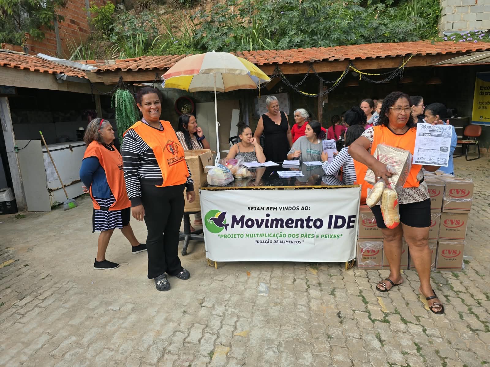 Voluntárias de colete laranja junto à mesa de atendimento do projeto, enquanto participantes aguardam a entrega de alimentos