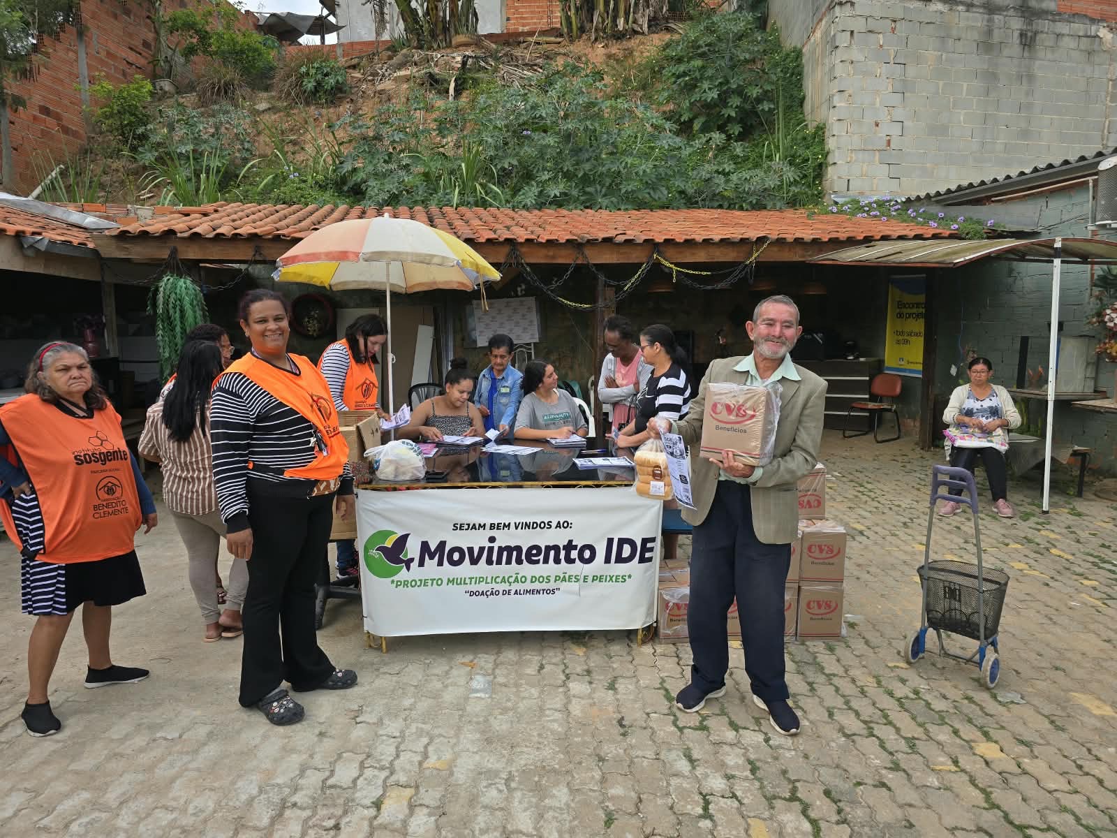Participante sorrindo com uma caixa de alimentos nas mãos ao lado da mesa do projeto e de voluntárias de colete laranja