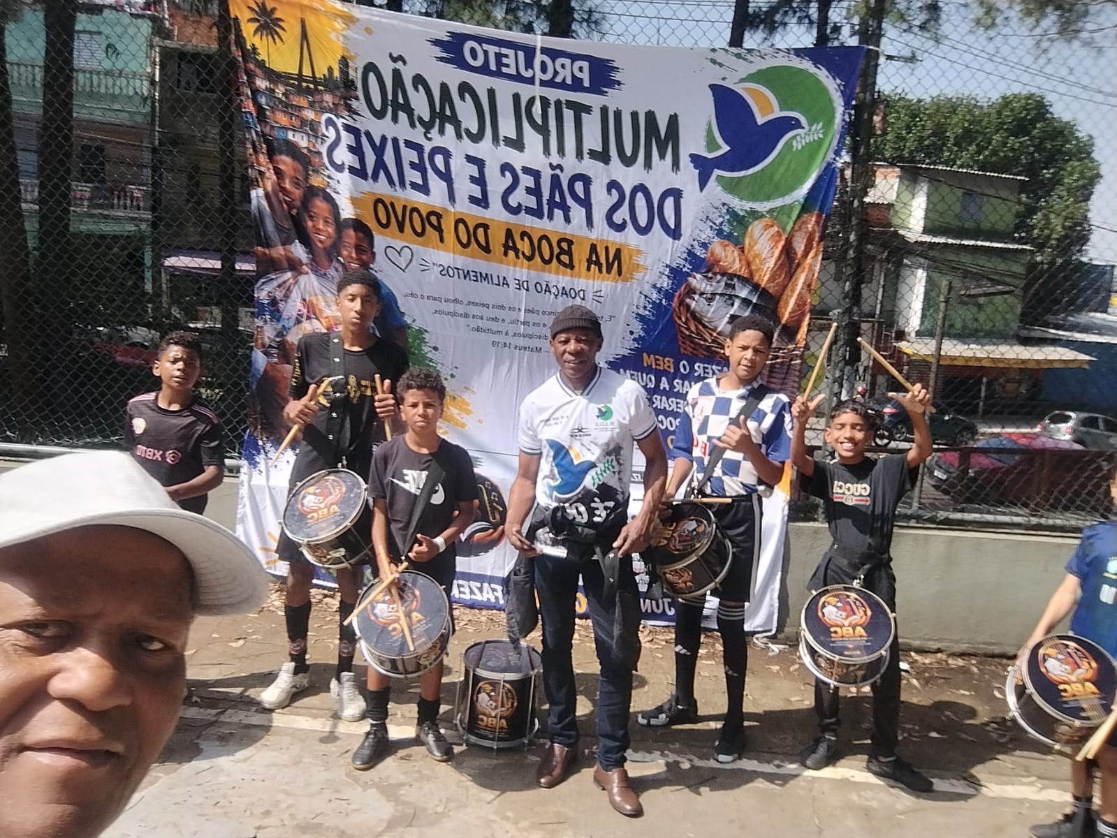 Jovens com caixas de percussão e educadores posando diante do banner do Projeto Multiplicação dos Pães e Peixes
