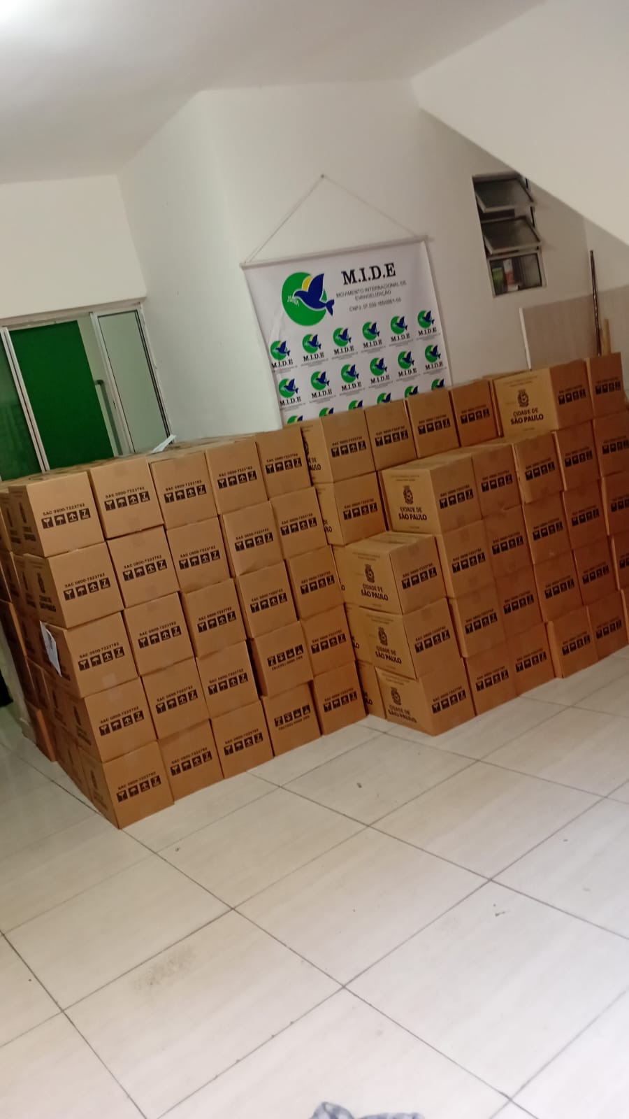 Grande quantidade de caixas de cestas básicas empilhadas em uma sala, diante de um banner do M.I.D.E